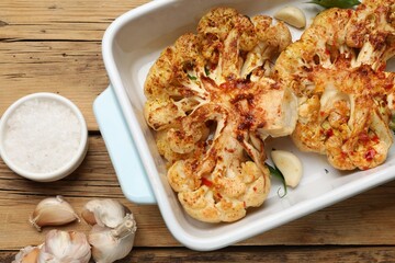 Tasty cauliflower steaks in baking dish and spices on wooden table, top view