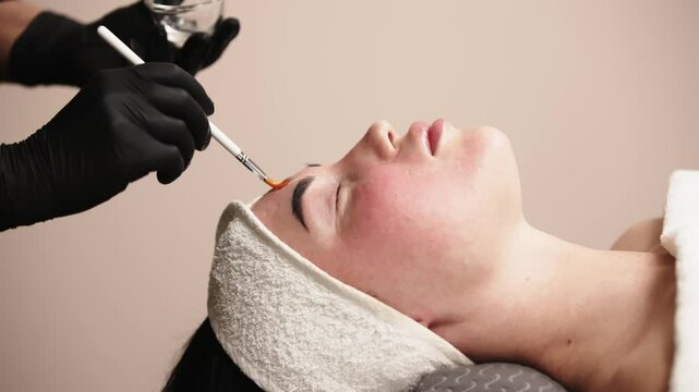 Close-up of a cosmetologist applying a cosmetic peeling mask to a client's face. The scene emphasizes the professional approach to skin care and the relaxing spa experience.