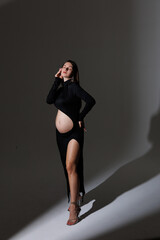 Fototapeta premium Beautiful young pregnant brunette girl. A woman waiting for a baby in a black dress, in the studio. Dress with a hole in the stomach, pretty pregnant belly.