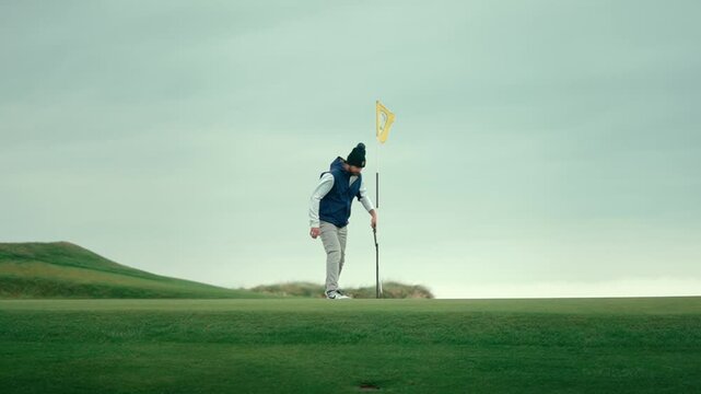 Male golfer approaches pin and reaches down to pick up ball after putting it into the hole on scenic Ireland golf course green, celebrates putt by throwing ball into the air