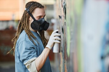 Artist wearing protective mask and gloves concentrating on creating colorful mural on city wall,...