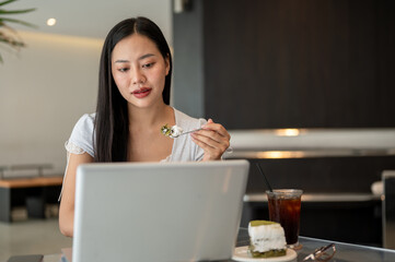 An attractive Asian woman is eating cake while focusing working on her laptop in a coffee shop.