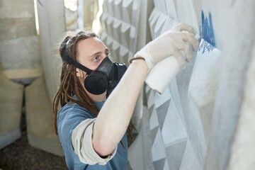 Graffiti artist painting detailed mural on outdoor wall, wearing respirator mask and protective gloves, focusing on unique patterns and textures in mural creation process © Seventyfour