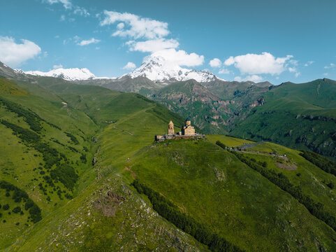 Aerial view of Gergeti Trinity Church surrounded by lush green mountains and snowy peaks, Stepantsminda, Georgia.