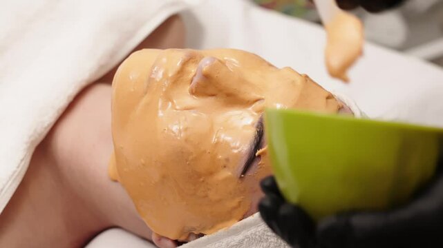 Close-up of a cosmetologist applying an alginate mask to a client's face with a spatula. The scene highlights professional facial skin care treatments in a beauty salon.