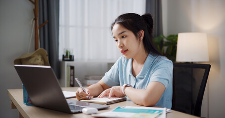 Portrait of Female freelancer sitting at desk taking notes on notebook while working on laptop in home office. Working from home, Remote working.