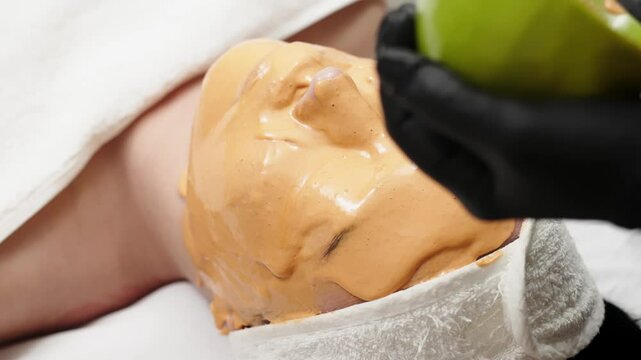 Close-up of a cosmetologist applying an alginate mask to a client's face with a spatula. The scene highlights professional facial skin care treatments in a beauty salon.