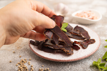 Armenian Basturma dried meat with hand on plate with pepper and herbs on brown concrete. Side view, selective focus
