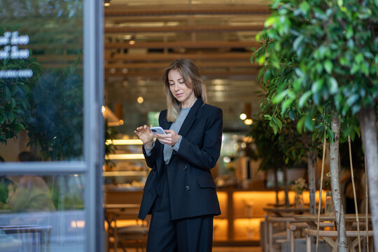 Businesswoman stands in cafe doorway using smartphone for chatting, surfing internet and managing business contracts. Woman cafe owner handles business correspondence and multitasks as manager.