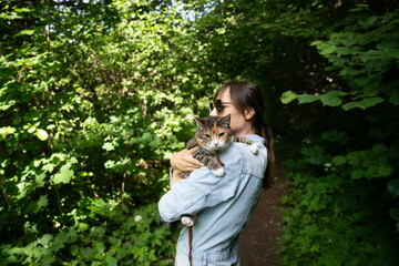 Obraz premium Woman pet sitter hugging embracing tabby cat strolling outdoors in summer day. Female owner and relaxed pet in hands wandering in public park, enjoying fresh air, warm sunny weather.
