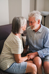 Elderly couple sitting on a cozy sofa, smiling and chatting with love and sweetness in their home. This heartwarming moment captures the essence of mature relationships, friendship, and the joy 