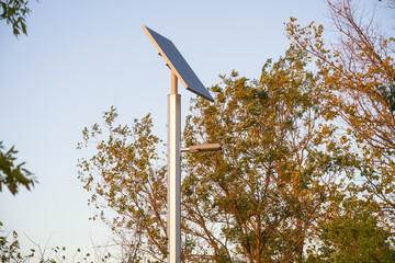 Close-up of a modern solar-powered street light illuminate a public park. The solar panel, perched atop a sleek metal pole, is surrounded by greenery as the light gently blends into the nature