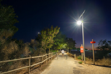 Rear view of a young single man jogging at night at the beginning of a footpath in a public park with modern solar powered street lights illuminating the country road. Spain, Europe