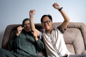 Down Syndrome Asian Boy And Mother in Hijab Celebrating Victory While Watching Sports Game at Home