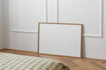 Horizontal thin picture frame mockup. Empty wooden frame poster mock-up template on oak wooden parquet floor. White wall background, stucco decor. Minimal Scandinavian interior, blurred bed sofa