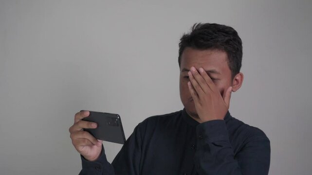 Southeast Asian man wearing dark blue shirt looking scared while looking at his phone while covering his face with his hands