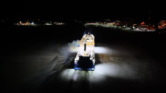 Hybrid roro ship moving through ice covered sea at night. Harbour lights ahead. Aerial stern view.