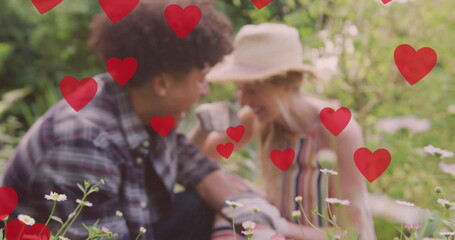 Image of red hearts over happy diverse couple gardening and laughing in sunny garden