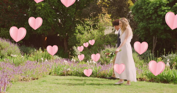 Image of pink hearts over happy diverse couple walking in sunny garden on wedding day