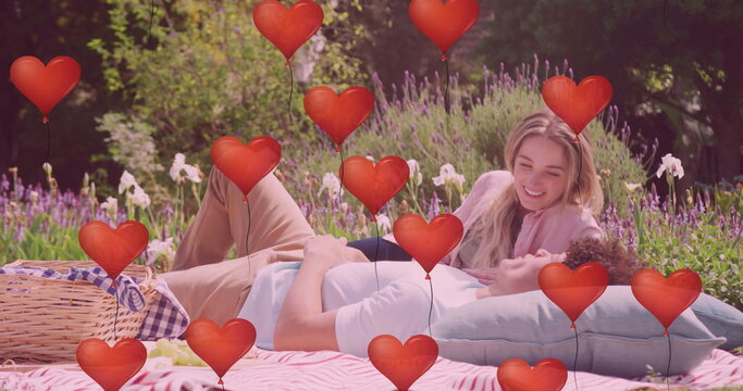 Image of red heart balloons over happy diverse couple relaxing at picnic in sunny garden