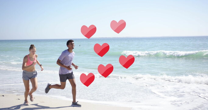 Image of red hearts over diverse couple running on beach