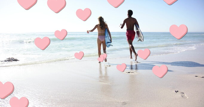 Image of pink hearts over diverse couple running with surfboards on beach