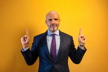A man in a suit and blue shirt is pointing upwards with his both hands, he is smiling. His background is yellow.