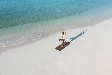 Aerial view of a woman practicing yoga on a mat with her shadow on the sand near gentle waves, Thoddoo, Maldives.