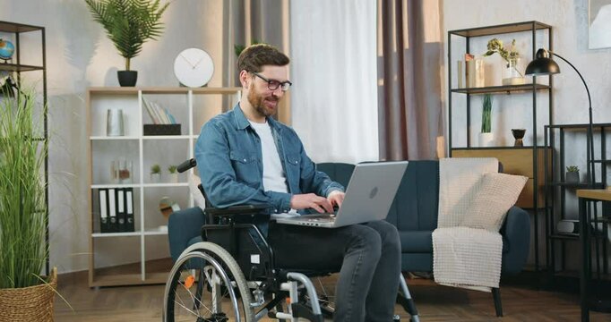 Disabled man in glasses working on computer laptop sitting in wheelchair after accident at home