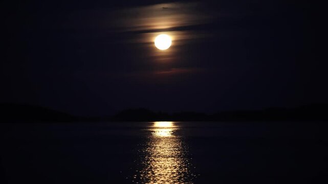 Dark night with full yellowish moon behind thin clouds, shimmering light reflecting on calm ocean surface with gentle ripples