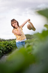 Obraz premium A young girl is standing in a field of green plants, holding a straw hat