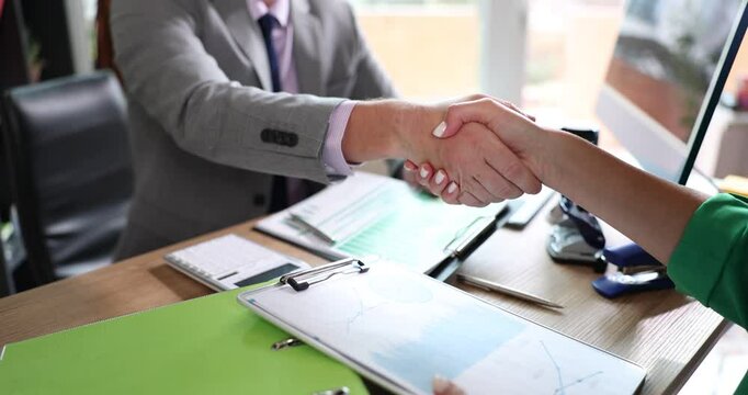 Two business people shake hands sitting at table with financial report charts. Partners shake hands after financial negotiations and are satisfied with result