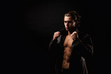 Perfect young man in unbuttoned leather jacket with abs muscle press posing in black studio shadow, strong look. Handsome guy fashion model with lovely muscular sexy torso, image. Copy ad text space