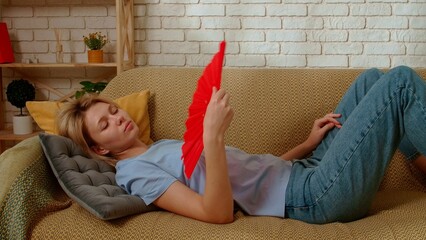Young millennial woman laying on the sofa in the living room waving fan, coping with summer heat,...