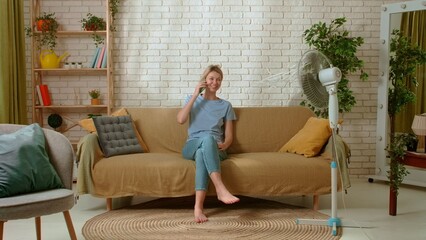 Obraz premium Young millennial woman sitting on the sofa in the living room talking on smartphone, electric air fan blows fresh air next to her. Stuffiness everyday life concept.