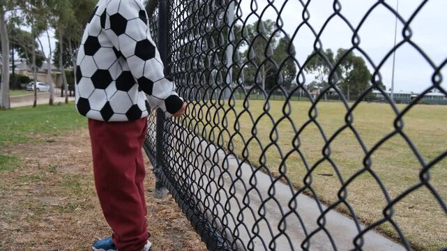 A young boy (5 year old) stands by the fence of a sports ground, occasionally bouncing his back against it as a repetitive behavior, seemingly lost in his world.