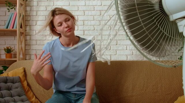 Young millennial woman sitting on the sofa in the living room relaxing under electric air fan blowing fresh air, close up shot. Stuffiness everyday life concept.