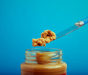 Peanut butter on a knife above a jar of peanut butter with a blue background.