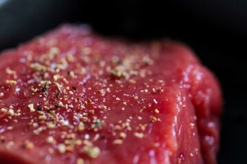 A close up portrait of the seasoning on a red raw steak being prepared or cooked in a frying pan. The delicious meat has salt and pepper on it for a perfect dinner or meal.