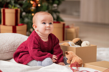 Fototapeta premium cheerful baby sits among gift boxes and toys next to a Christmas tree.
