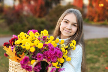 Naklejka premium Portrait of a pretty teen girl in a cozy white sweater with bouquet of autumn flowers in basket in autumn garden. Outdoor portrait of smile girl in knitted clothes holding chrysanthemums on backyard 