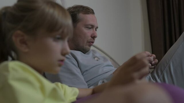 Dad and daughter sit at home and play video games on a mobile phone
