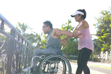 Asian disabled person sits in a wheelchair with a friend pushing in a city park and exercises for good health, having fun in the gentle spring sunshine.