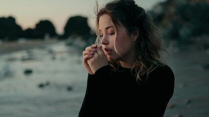 A woman stands on a chilly beach, hands clasped together as she tries to warm up. The ocean waves create a serene background, capturing a moment of solitude and reflection.