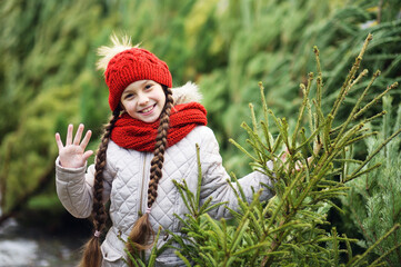 little girl with pigtails in a red knitted hat and scarf smiles on the background of the Christmas tree market and chooses a Christmas tree