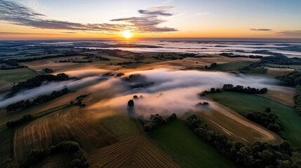 Obraz premium Aerial view of a sunrise over a foggy landscape with rolling hills, fields, and scattered trees. The light mist creates a serene atmosphere with vibrant colors in the sky.