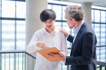 A young asian man standing and conversation with advisor teacher in a building,professor giving advice and university student with clipboard, support and touching his shoulder