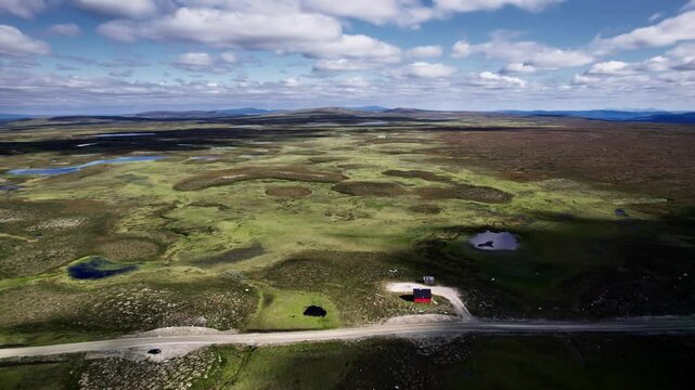 Ascending drone reveals a red building beside a winding road in the vast, open high fell of Flatruet, showcasing waterholes and beautiful moorland in Sweden.
