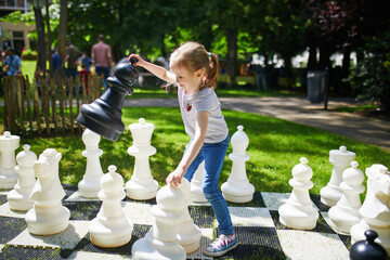 Adorable preschooler girl playing chess with big chess pieces outdoors on a sunny summer day.
