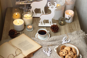 Various Christmas decorations, lit candles, cup of tea or coffee, cookies, books, presents and soft blanket on the table. Holiday hygge at home. Selective focus.